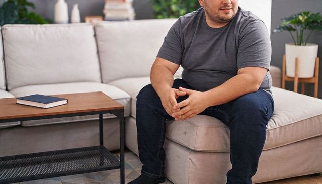 Man sitting on beige sofa wearing gray t-shirt and jeans clasping hands in home interior