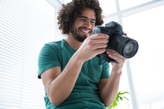 Photographer with curly hair smiling while reviewing photos on his digital camera in a well-lit studio. Ideal for use in articles or advertisements related to photography, creative professions, technology, and hobbies. Can also be used for promoting photography courses, camera equipment, or studio setups.
