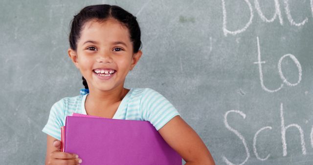 A young girl of diverse ethnicity smiles brightly while holding a purple folder in front of a chalkboard with the words Back to School written on it, with copy space. Her cheerful expression and the classroom setting suggest excitement for a new school year.