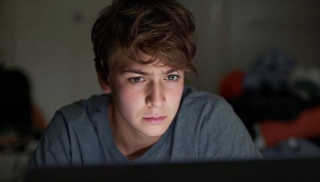 Teenage boy leaning toward laptop in dim bedroom focusing intently on screen