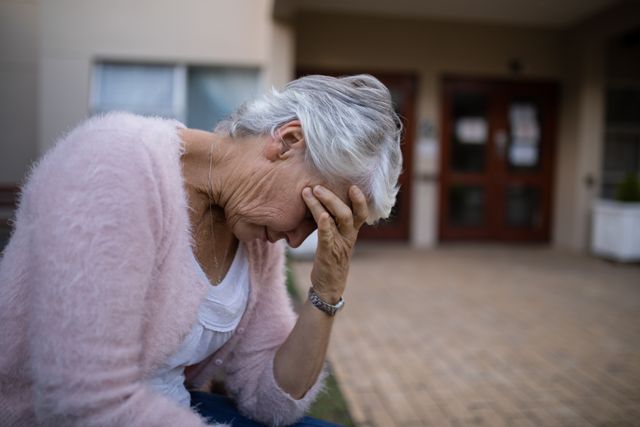 Side view of depressed senior woman sitting on bench with head in hand against nursing home