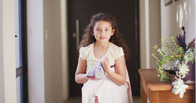 Young girl with curly hair carrying folded clothes in a sunlit hallway. The scene represents home responsibilities, helping out, and family life. Perfect for use in content related to childhood, family, home chores, educational materials on responsibility, and promoting helpful behavior in children.