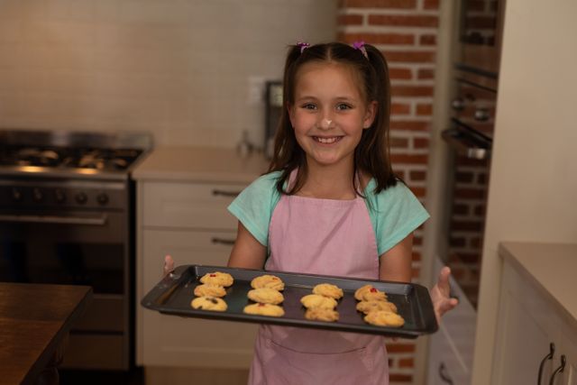 A caucasian girl wearing an apron in the kitchen holding a tray of cookies up with a smile on her face. a smudge of flour is on her nose.