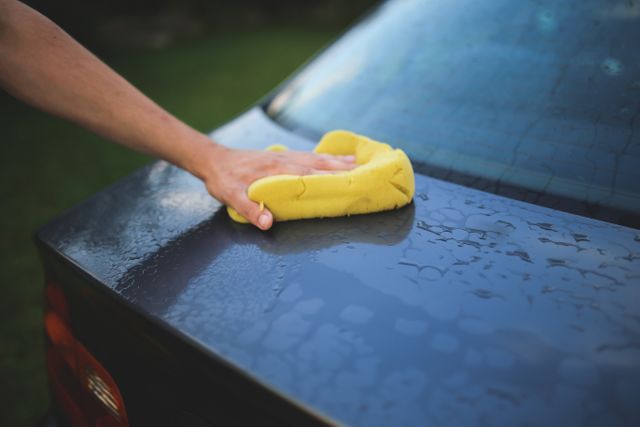 Hand cleaning car with yellow sponge in outdoor setting. Suitable for car care products, car washing services, maintenance tips, advertisements for car detailing products, and DIY car wash guides.