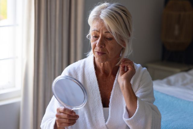 Mature caucasian woman in bathrobe looking at herself in hand mirror in bedroom, copy space. Self care, health, beauty and senior lifestyle concept.