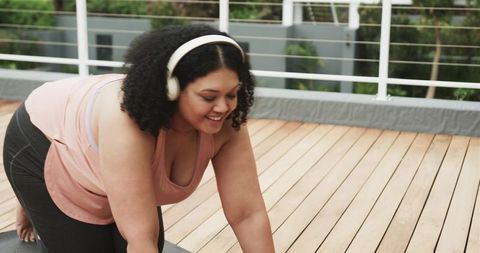 Curly-haired woman practicing yoga on balcony deck with headphones and workout clothes