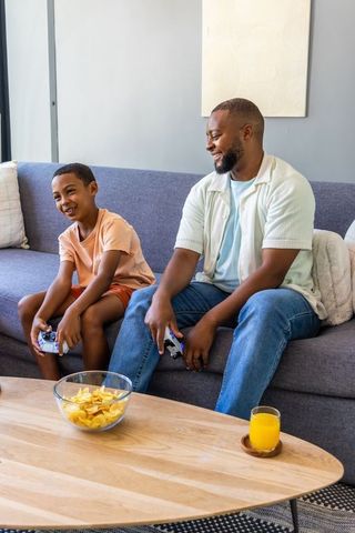 African american father and son playing video games on sofa sharing snacks and laughing together