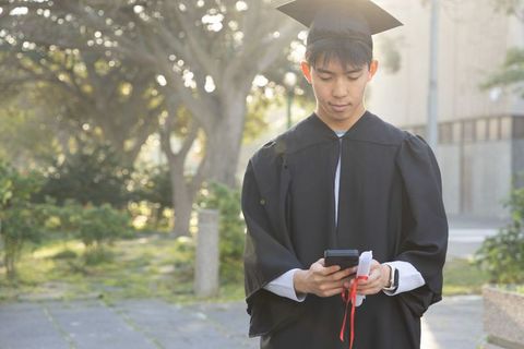 Asian graduate checking smartphone while holding rolled diploma on sunlit campus courtyard