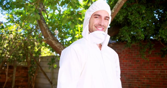 A young man is smiling and wearing protective gear including a hooded suit, mask, and gloves in an outdoor setting under a tree. Ideal for content related to environmental safety, occupational health, personal protective equipment advertising, and articles about working in nature.