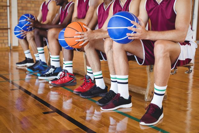 Basketball players sitting on bench holding basketballs in indoor court. Ideal for sports team promotions, basketball training programs, athletic footwear advertisements, and articles on teamwork and preparation in sports.