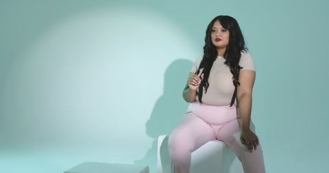 Curvy woman sitting on white cube wearing black suspenders holding slim object in pastel studio