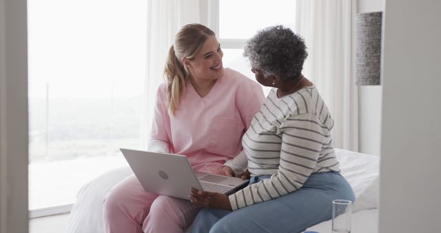 Senior woman sitting on bed using laptop with caregiver. Caregiver and senior enjoying technology together. Ideal for content about elder care, healthcare at home, use of technology in healthcare, and caregiving.