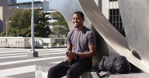 African American man smiling on urban plaza ledge wearing sportswear holding water bottle