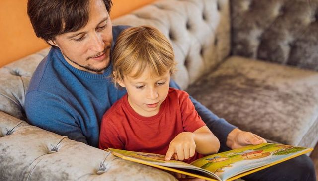 Father reading picture book with young son on cozy tufted gray sofa, bonding moment