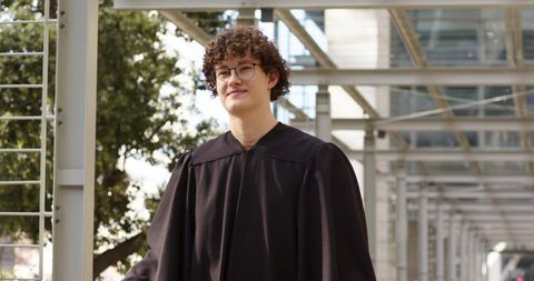 Young graduate wearing black gown and glasses in modern campus colonnade