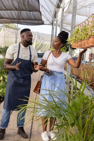 African american nursery staff discussing hanging plants with customer in greenhouse