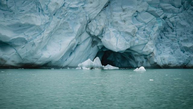Massive glacial ice cave meeting calm teal fjord water and scattered floating icebergs in arctic sea