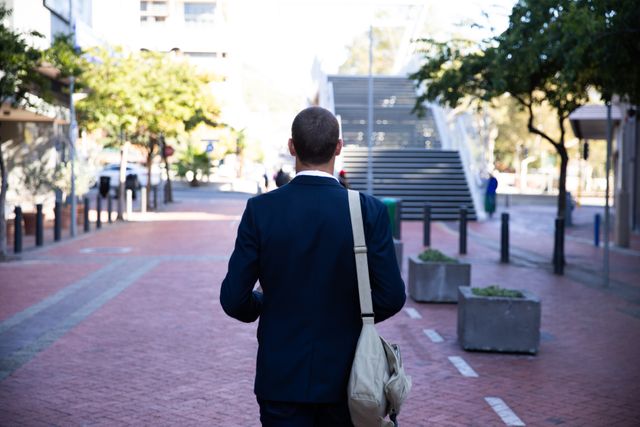 Rear view of elegant Caucasian businessman with a bag on his shoulder wearing a suit walking down the city street. Digital Nomad on the go.