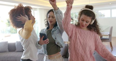 Diverse friends dancing and singing at home wearing headphones and sharing smartphone
