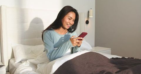Smiling asian woman using smartphone while sitting up in bright modern bedroom