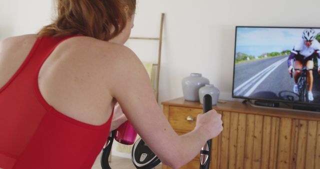 This image shows a woman engaging in a virtual cycling program from the comfort of her home. She is using a stationary bike while watching a cycling race on the television. This can be used for promoting home fitness programs, virtual exercise solutions, or cycling equipment.