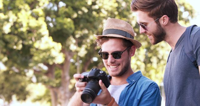 Two friends outdoors on sunny day, smiling and reviewing photos on camera. Used for themes related to friendship, travel, technology in leisure time, and outdoor activities.