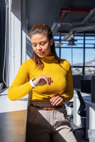 Checking smartwatch in sunlit modern office, woman leaning on windowsill