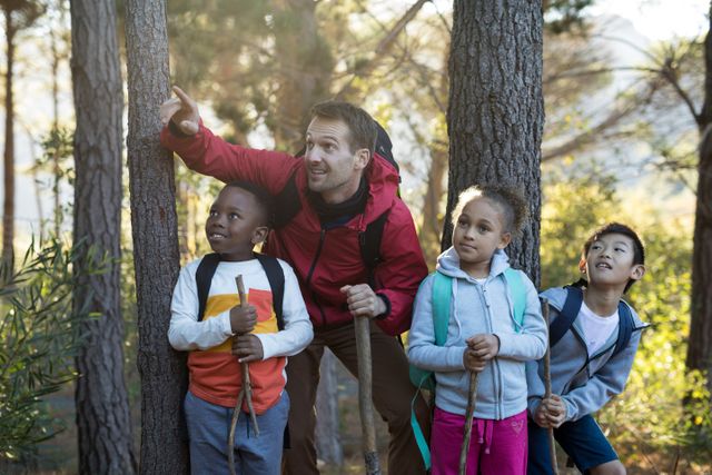 Teacher guiding diverse group of children on a nature hike in a forest on a sunny day. Ideal for use in educational materials, outdoor adventure promotions, and articles on childhood development and environmental education.