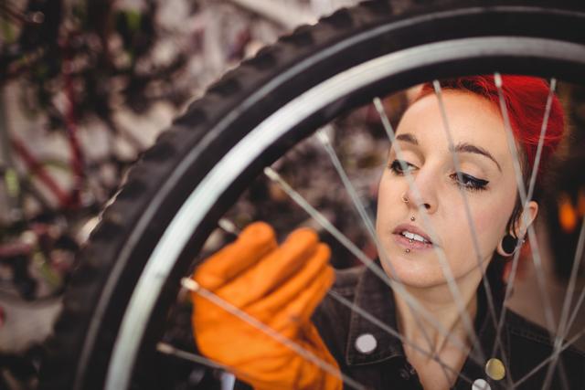 Young female mechanic with red hair closely inspecting a bicycle wheel in a workshop setting, wearing orange gloves for protection. This image can be used for promoting bike repair services, technical workshops, or bicycle maintenance instructions.
