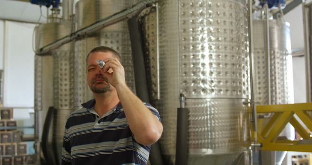 Man in striped shirt using instrument to inspect product quality at modern industrial brewery. Stainless steel tanks and machinery in background highlight industrial setting. Useful for content on brewing processes, quality control measures, industrial production, and beverage industry operations.