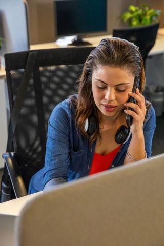 Professional woman taking work call at office desk wearing headphones and using computer