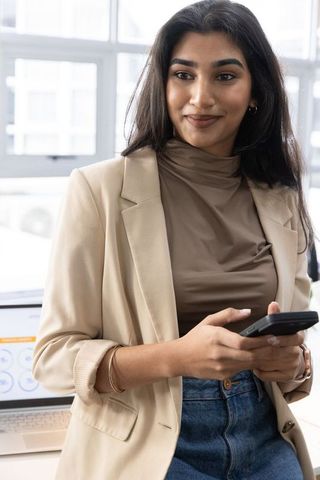 Indian professional woman using smartphone in modern office wearing beige blazer and jeans
