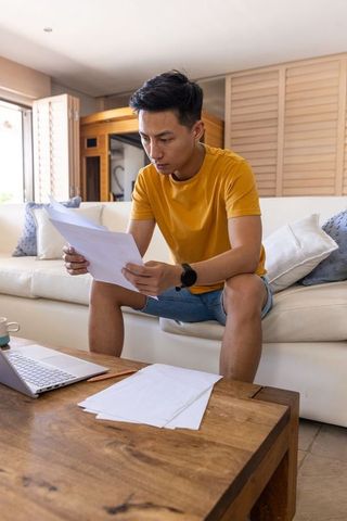 Asian man reading documents on sofa with laptop, working from home in bright living room