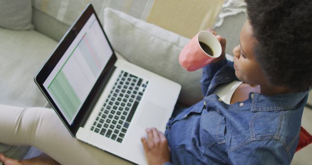 Young woman, wearing casual clothes, sitting on a couch while working on her laptop and holding a cup of coffee. This image can be used in articles about remote work, freelancing, work-life balance, or productivity tips. Perfect for blogs and websites focusing on modern work environments or inspirational content about achieving goals.