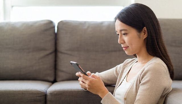 Woman sitting on sofa using smartphone, scrolling messages in calm modern living room