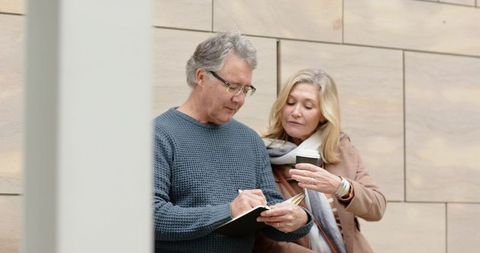 Mature couple planning travel outdoors, man writing notes while woman holding coffee