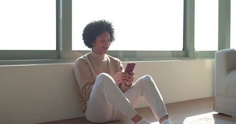 Sunlit african american man sitting on floor using smartphone in minimal apartment