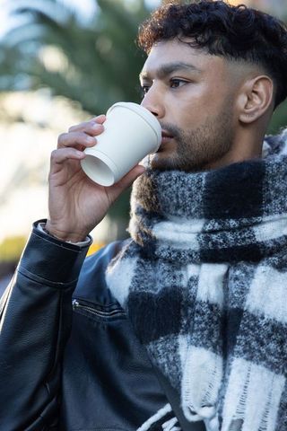 Man sipping coffee from disposable cup wearing leather jacket and plaid scarf outdoors