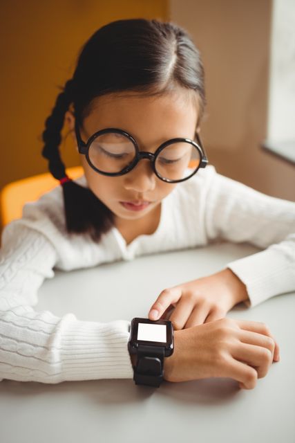 Young girl with braided hair and glasses wearing a smart watch while sitting at a desk in a classroom. Ideal for use in educational materials, technology in education promotions, or modern classroom settings.