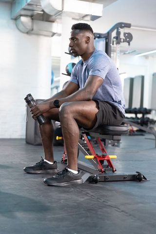 African American man sitting on adjustable bench holding water bottle resting between gym sets