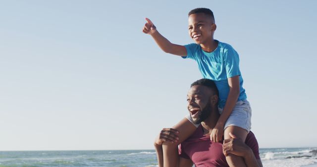 Father and son enjoying a fun day at the beach. Son is sitting on his father's shoulders, both smiling and pointing at something exciting in the distance. Ideal for depicting family bonding, joyful childhood moments, summer vacations, and outdoor activities.
