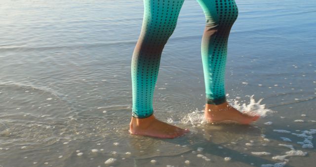 A woman is seen from the knee down wearing teal leggings while walking in shallow ocean water at the beach. Ideal for concepts of active lifestyle, summer activities, fitness, outdoor enjoyment, coastal vacations, and nature relaxation.