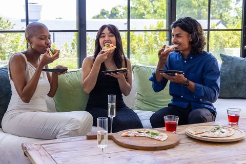 Diverse friends sharing pizza on sofa during casual home gathering with garden view