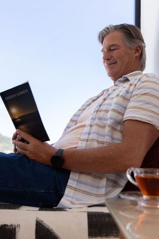 Mature man relaxing with book and tea by window, coastal view, striped shirt, sunset brochure
