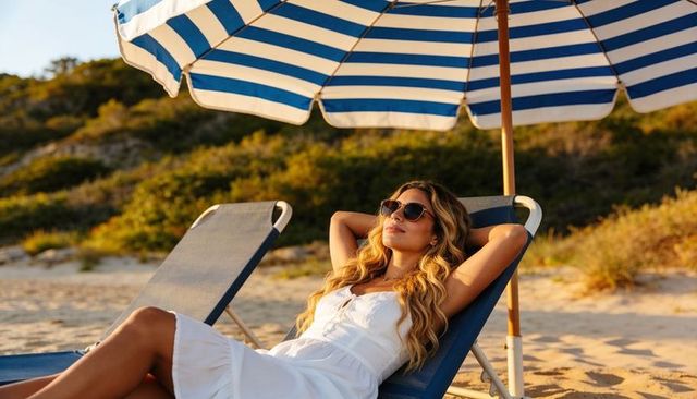 Young woman relaxing on lounge chair under blue-striped umbrella on golden sunlit beach