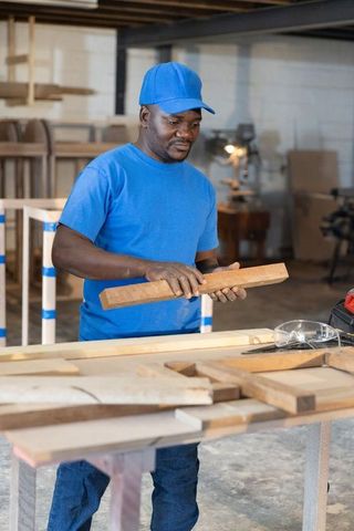 African american craftsman inspecting wooden plank in carpentry workshop wearing blue cap