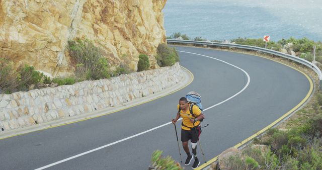 African american man with prosthetic leg trekking with walking poles and backpack on coastal road. Long distance walking, fitness, challenge, disability and healthy outdoor lifestyle.