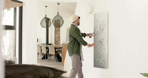 Indian man adjusting textured wall art in modern open-plan home with natural light
