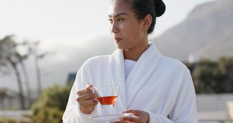 Asian woman wearing white bathrobe sipping tea on terrace with mountain view