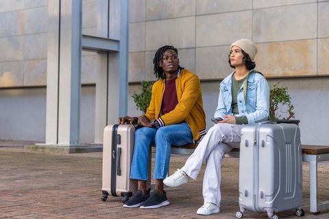Waiting with luggage at urban plaza, diverse travelers sitting on bench using smartphone